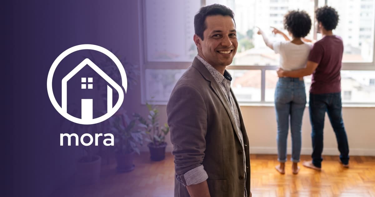 Corretor sorrindo em um apartamento enquanto um casal observa a vista pela janela, ao lado da marca Mora